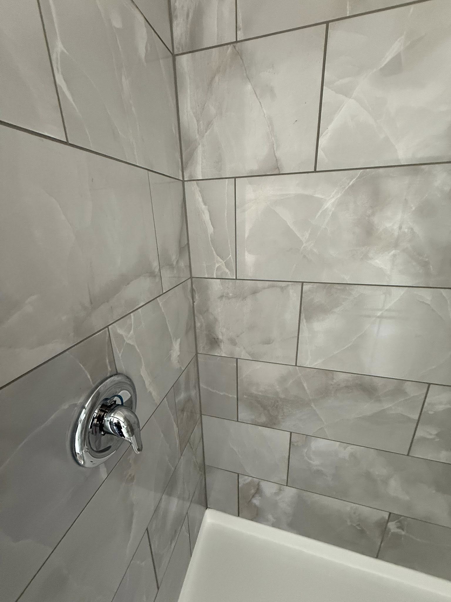 Close-up of a shower corner with glossy marble-patterned tiles in gray and beige tones. A shiny chrome shower handle is mounted on the left wall.