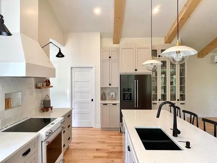 Modern kitchen with white cabinets, black appliances, and a central island with a sink. Pendant lights hang from wooden beams, creating a warm, inviting space.