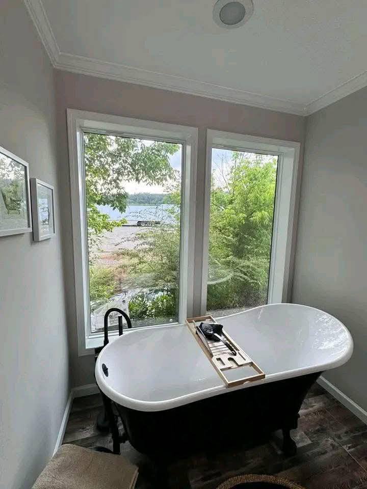 A white freestanding bathtub sits in front of two large windows with a view of leafy green trees and a distant lake, creating a serene atmosphere.