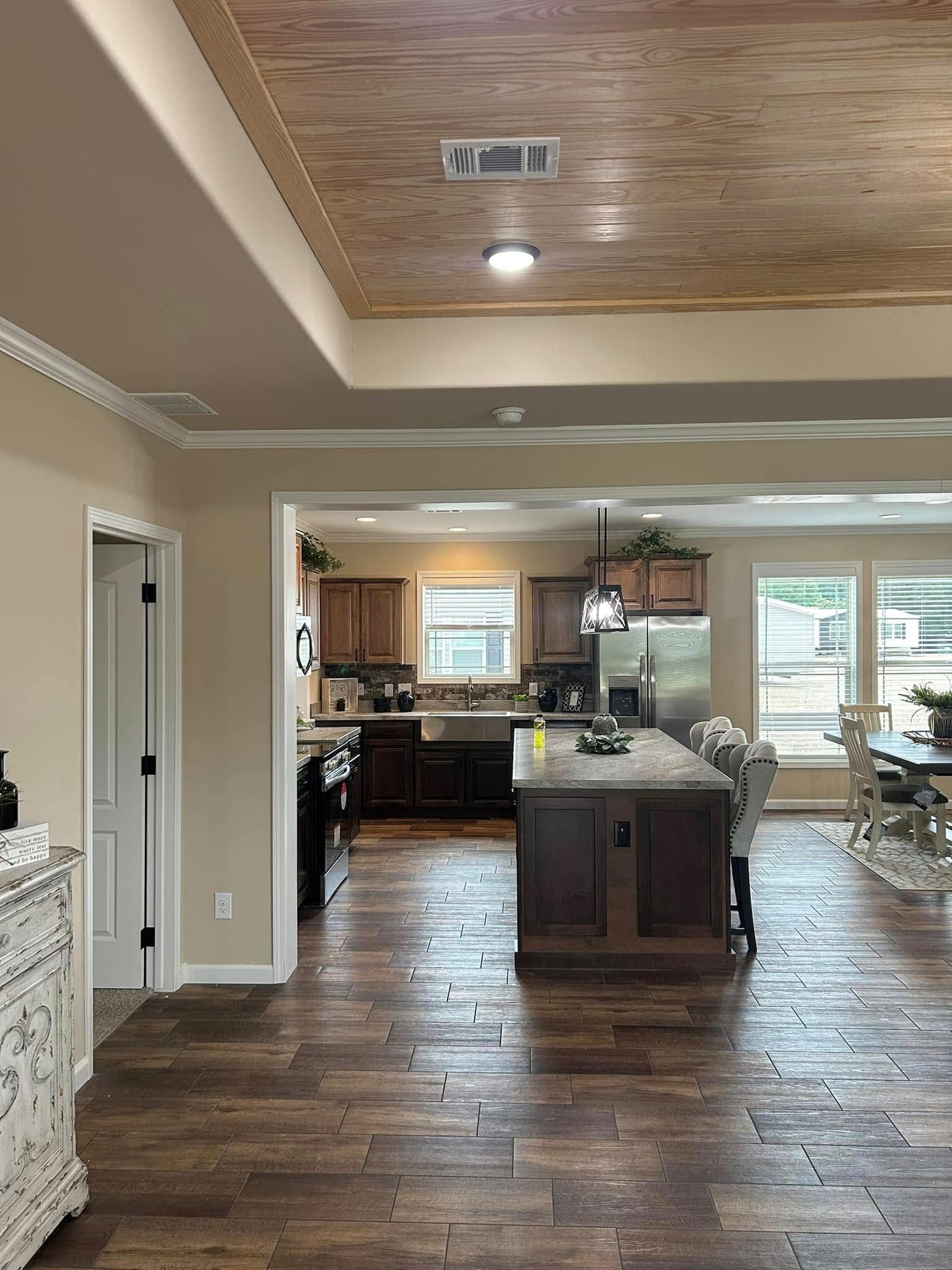 Modern kitchen and dining area with wooden floors, a central island, dark cabinets, and large windows. Bright, inviting atmosphere, and natural light.