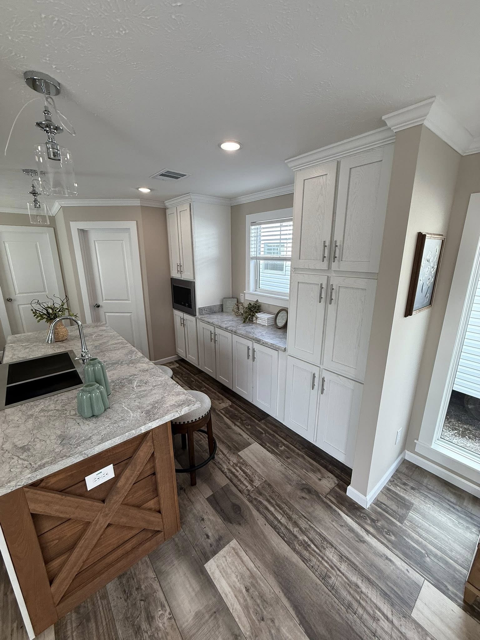 Bright modern kitchen with light wood flooring and beige walls, featuring white cabinets, a marble countertop, and teal vases on the island. Warm, inviting feel.