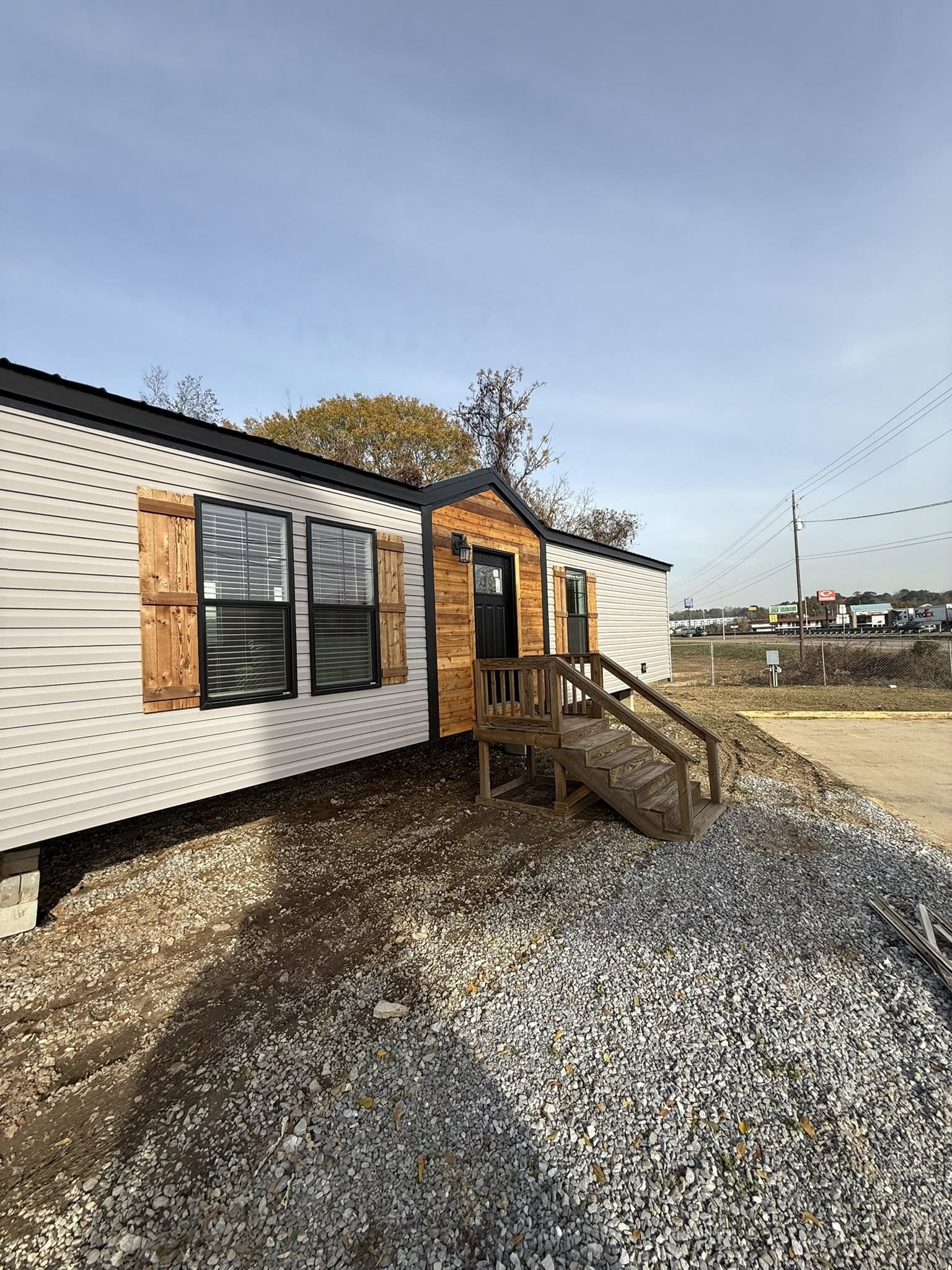 Single-story modular home with white siding and wooden accents. Wooden shutters and stairs lead to a black door. Gravel yard, clear sky. Industrial area in background.