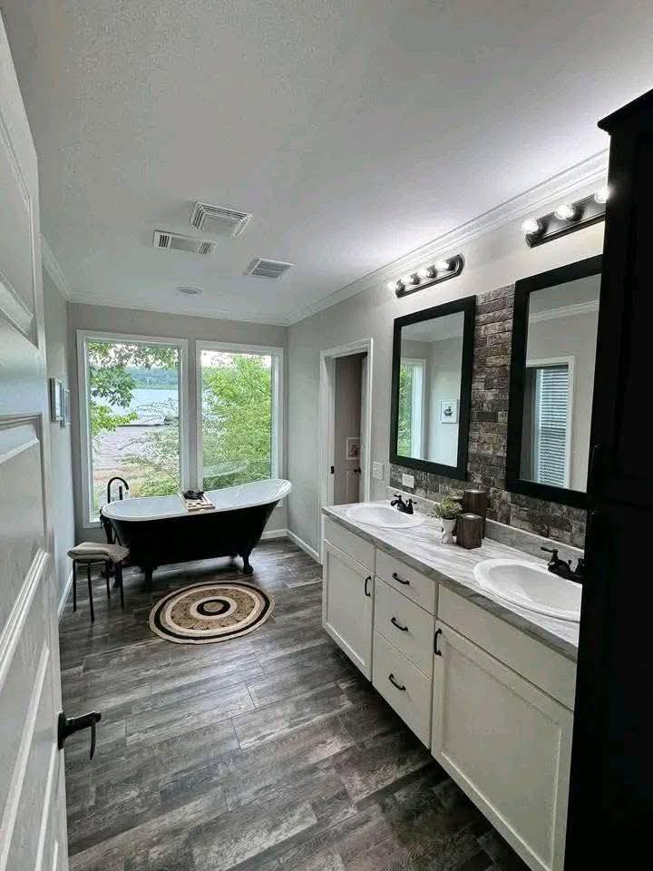 Elegant bathroom with dark freestanding tub, large windows overlooking greenery, double vanity with mirrors, and rustic wood floor. Modern, serene vibe.