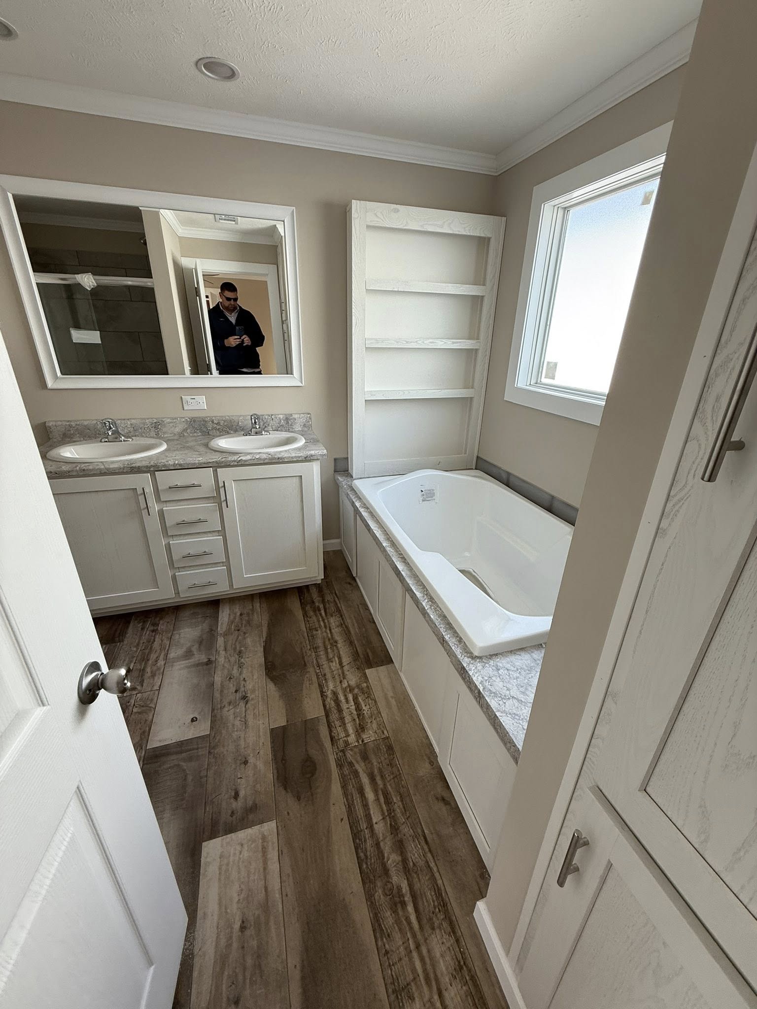 Modern bathroom with dual sinks, spacious bathtub, and light wood flooring. A person is partially visible in the mirror, adding depth to the scene.