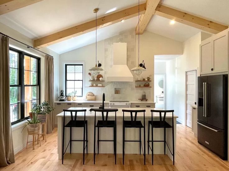 Bright kitchen with vaulted ceiling and wooden beams. Features a white island with black bar stools, pendant lights, large windows, and modern appliances. Cozy and inviting atmosphere.