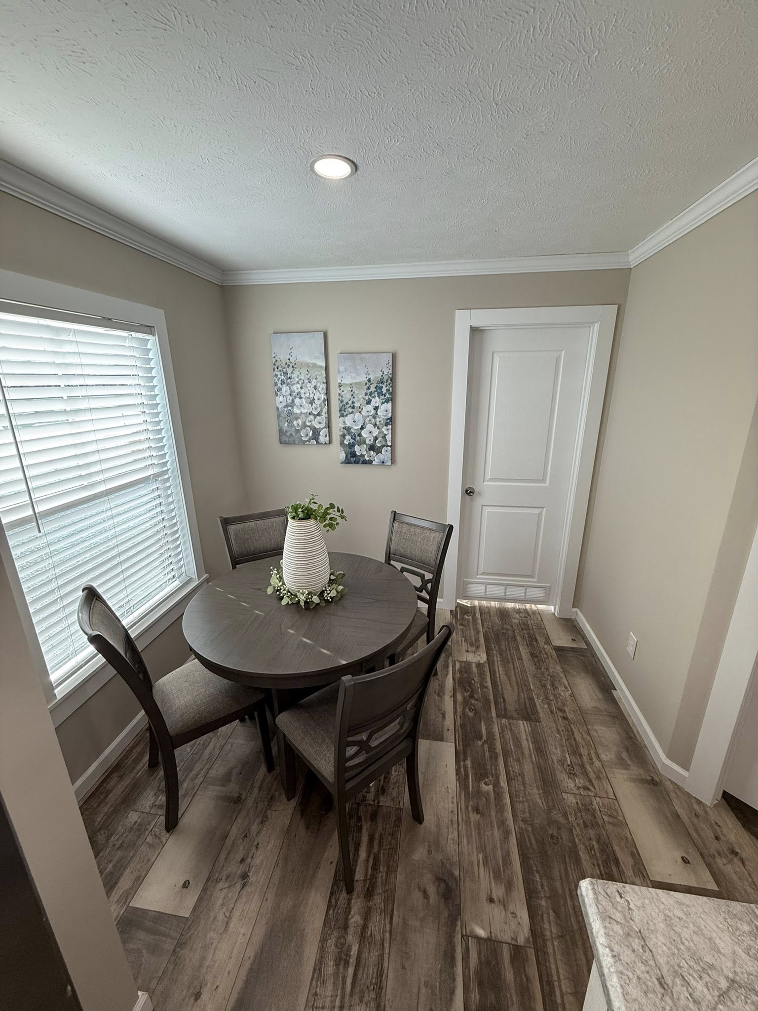 Cozy dining area with a round wooden table, four chairs, and a white vase centerpiece. Two floral paintings adorn beige walls; sunlight filters through blinds.