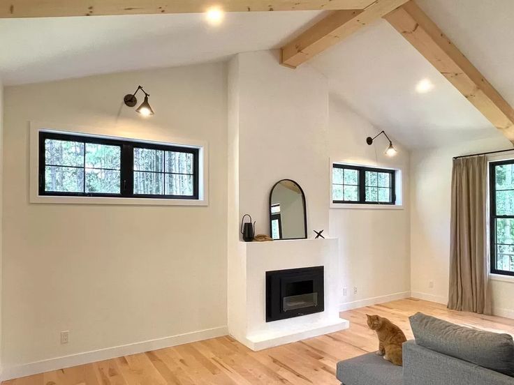 Modern living room with a white fireplace, adjacent windows, and exposed beams. A cat sits on a gray sofa, adding a cozy touch to the minimalist design.