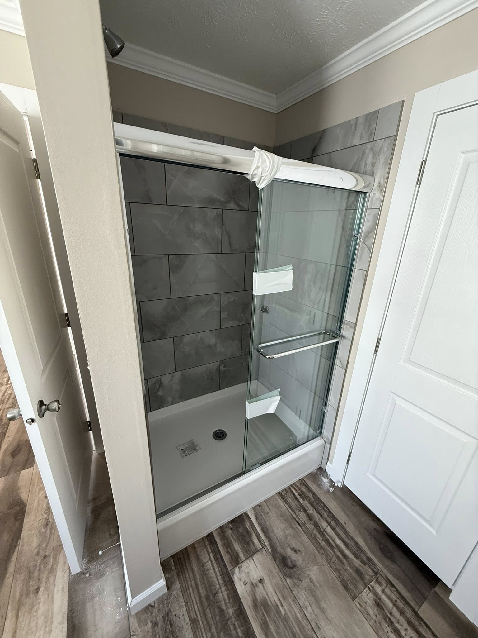 A small bathroom with wood-look vinyl flooring, featuring a walk-in shower with glass sliding doors and gray tiled walls. The atmosphere is clean and modern.