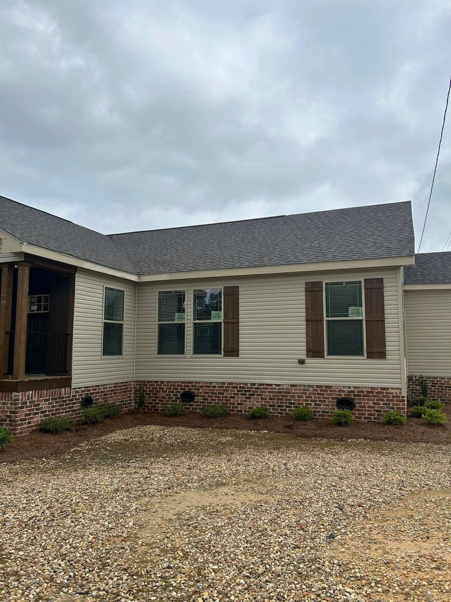 Single-story house with beige siding and brick foundation, featuring two large windows with wooden shutters. Overcast sky adds a calm tone.