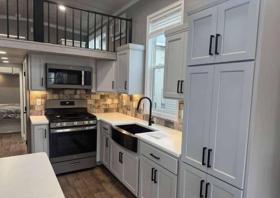 Modern kitchen with gray cabinets, a black farmhouse sink, and stainless steel appliances. Natural light from a window and a loft space above.