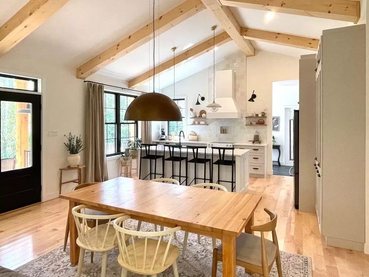 Bright modern kitchen with wooden beams on a high ceiling, a large wooden dining table, white chairs, and a central island with stools. Warm, inviting ambiance.