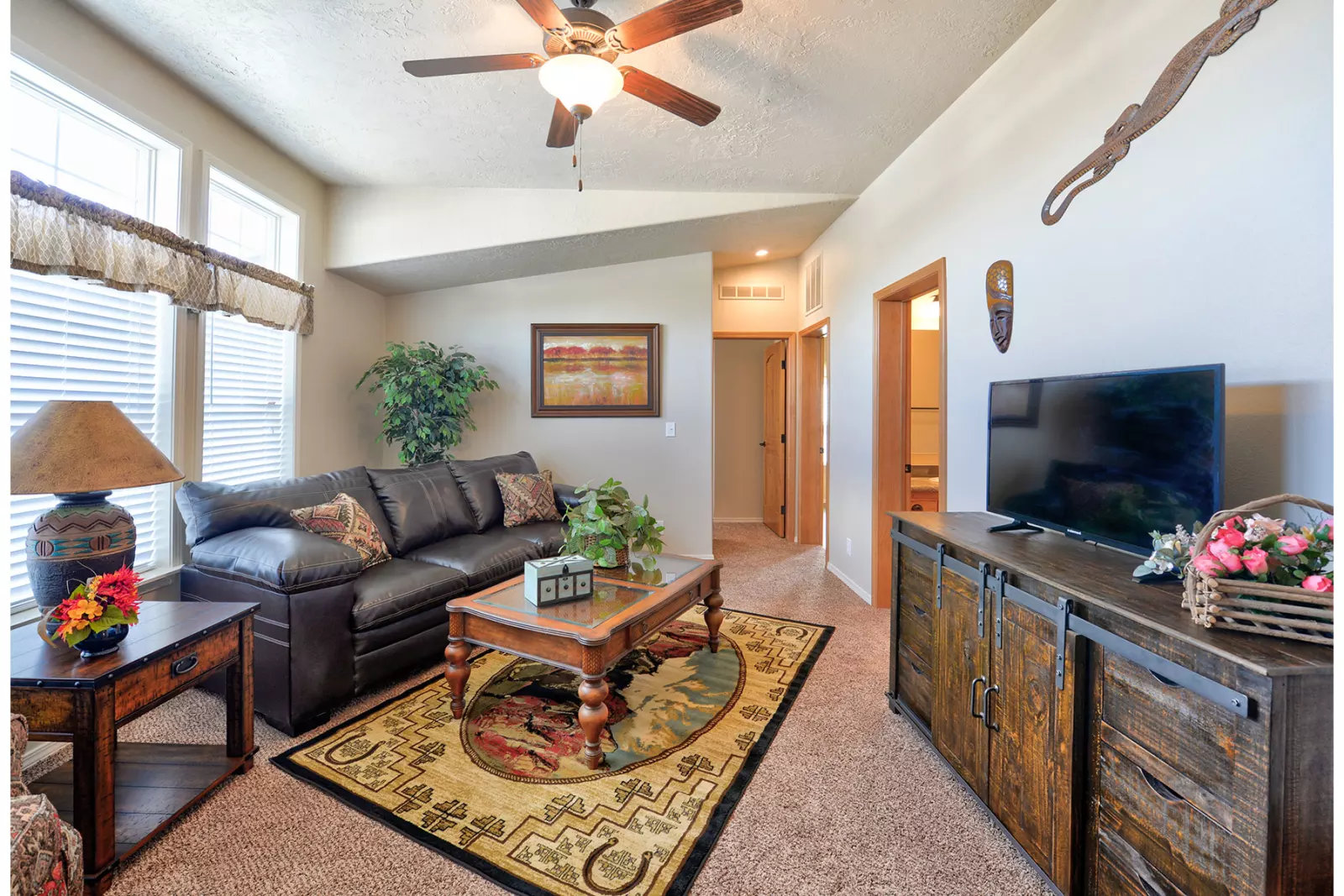 Cozy living room with brown leather sofa, colorful rug, and wooden furniture. A ceiling fan, large windows, and decorative plants add warmth.