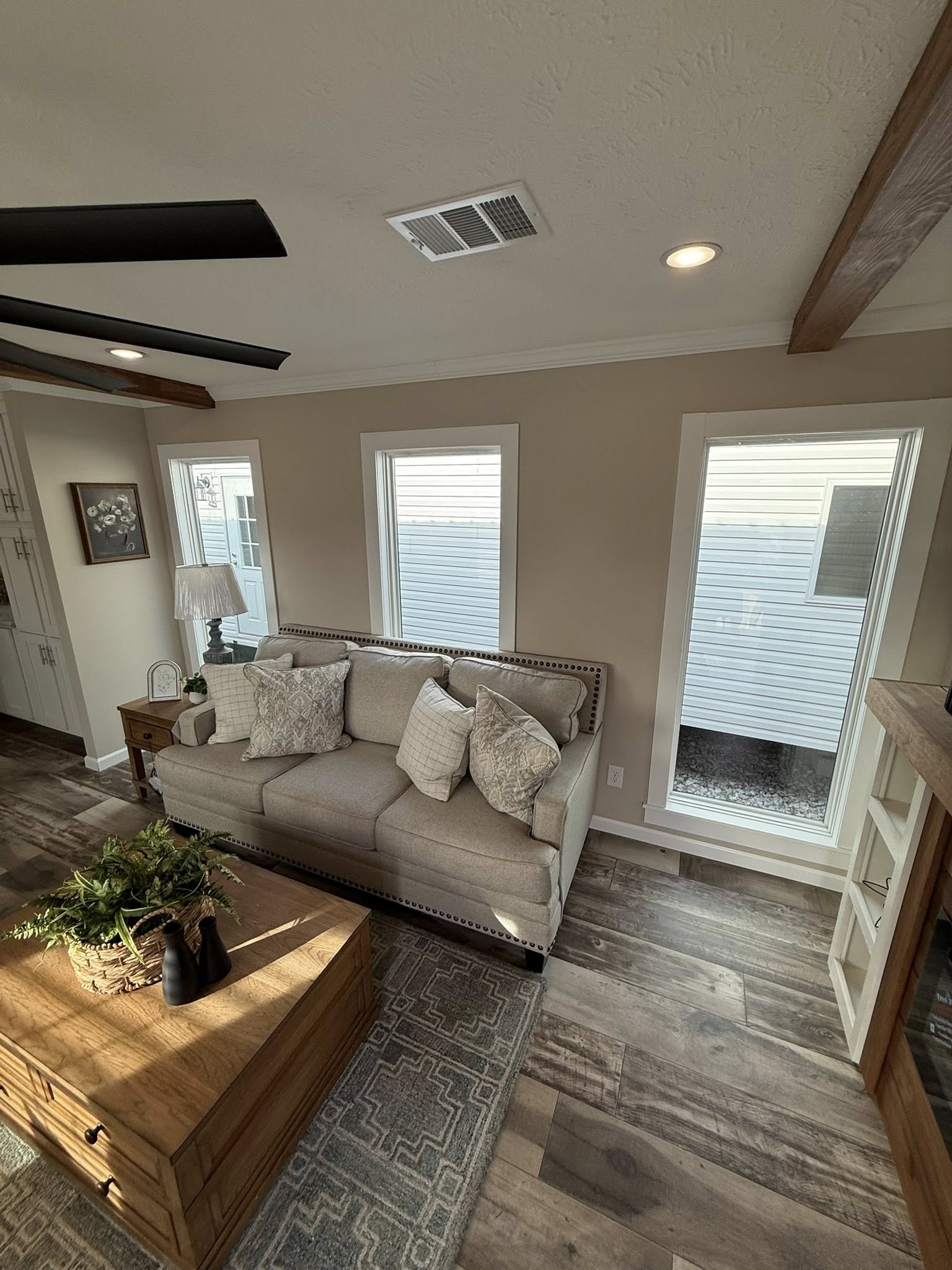 A cozy living room with a beige sofa adorned with patterned cushions, framed by three tall windows. Warm wooden floors and a rustic coffee table add a welcoming tone.