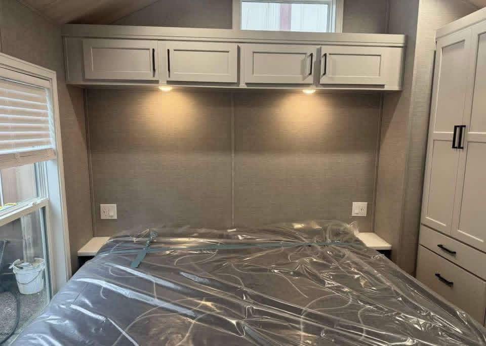 A cozy bedroom featuring a bed with plastic-covered mattress, light gray overhead cabinets with built-in lighting, and a window with blinds, conveying a clean, modern feel.