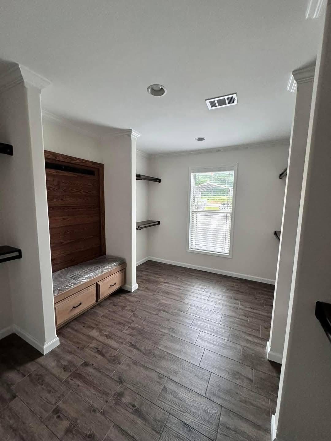 A small, empty room with dark wood flooring, a large window with blinds, and a built-in wooden bench and shelves along the walls. The atmosphere is clean and minimalist.