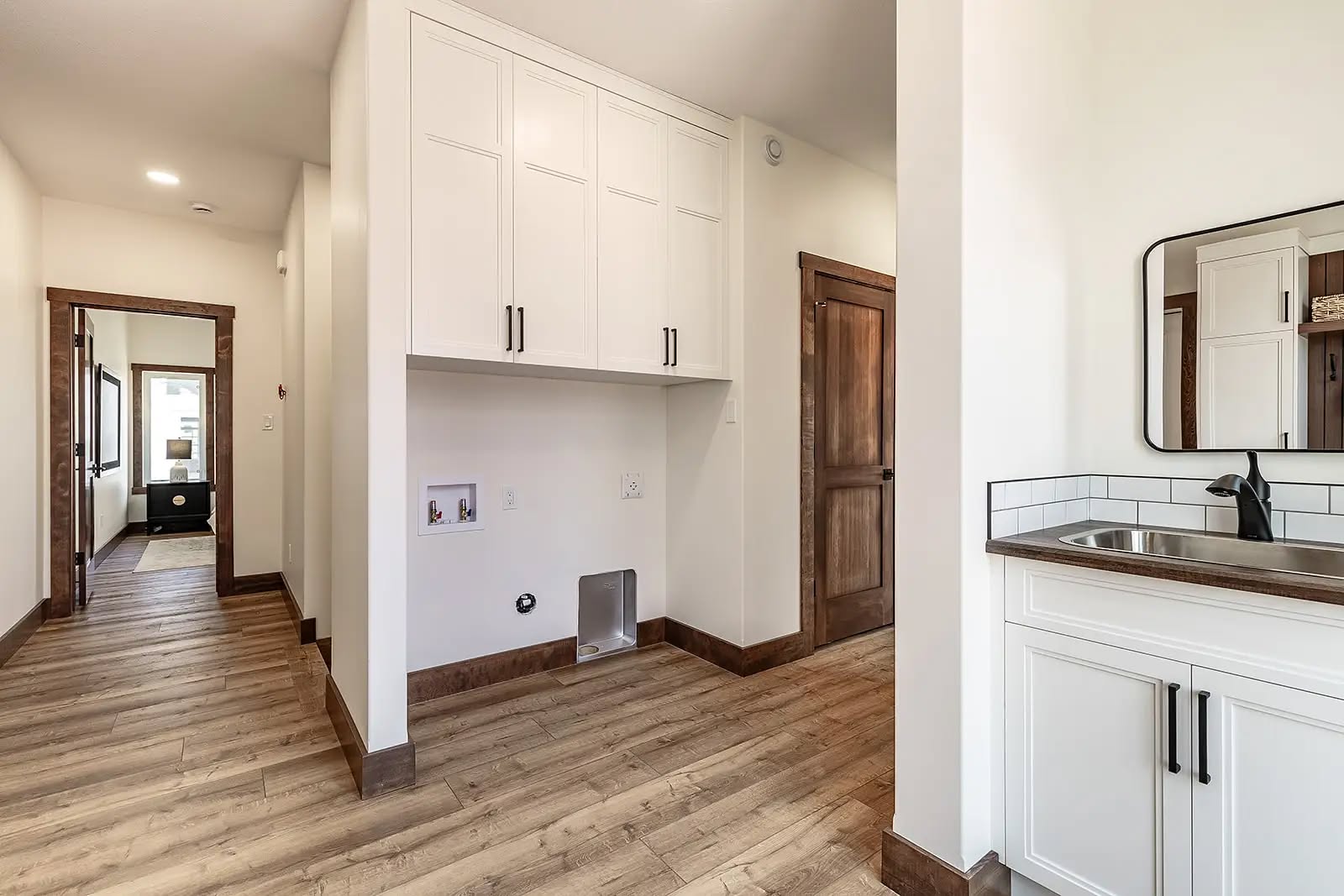 A modern laundry room features white cabinets, a brown wooden door, and a sleek sink with a mirror. The light wooden floor adds warmth.