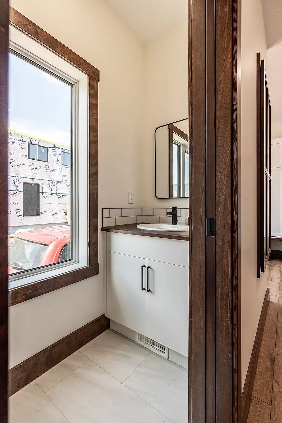 A narrow bathroom with a white vanity, black faucet, and rectangular mirror. A large window lets in natural light. The decor is modern and minimalist.
