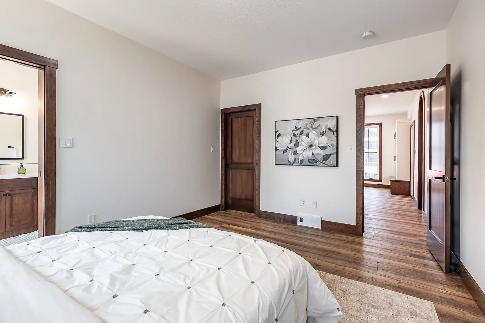 Minimalist bedroom with wooden accents and soft lighting. A bed with a white comforter and green throw faces a hallway and bathroom. A floral painting adorns the wall.