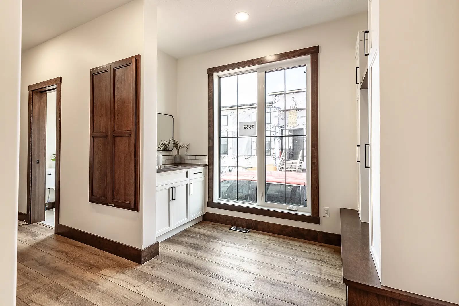 Bright hallway with wood flooring and a large window. Features a white cabinet with plants to the left and wooden trim accents, creating a cozy atmosphere.
