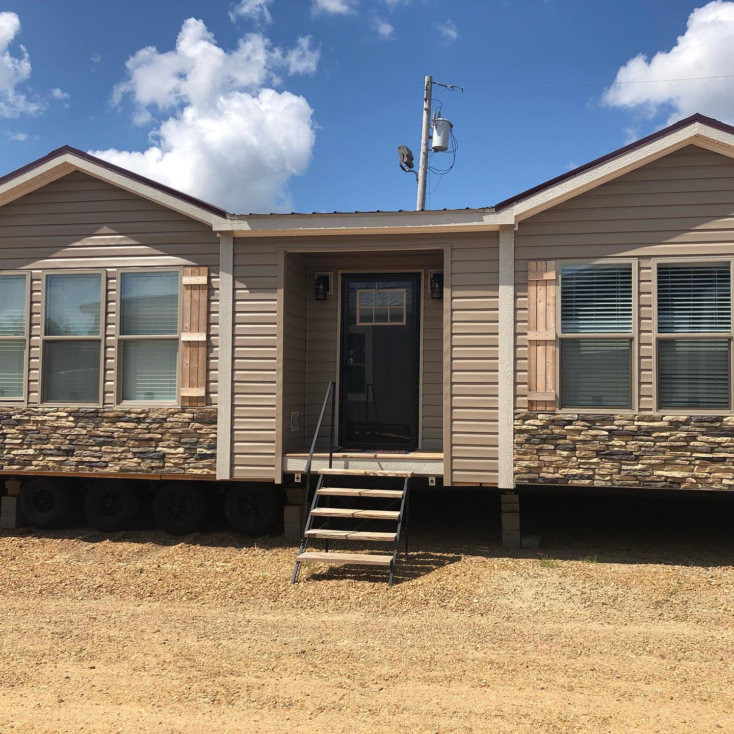 Single-story modular home with beige siding and stone accents, set on a dry, bare landscape. Stairs lead to a dark front door under a bright, cloudy sky.