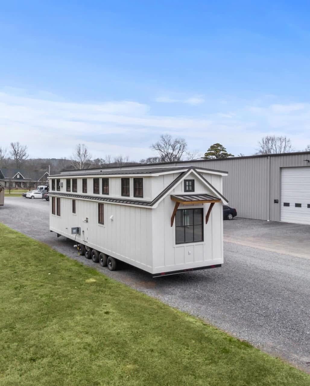 A white, modern tiny house on wheels with a gabled roof is parked on a gravel driveway. It's beside a large industrial building and a grassy area under a clear blue sky.