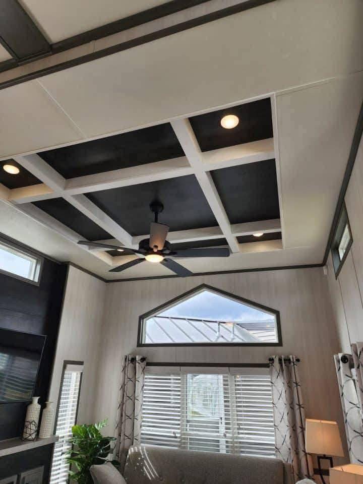 Modern living room with a coffered ceiling featuring recessed lights and a ceiling fan. Large windows with patterned curtains allow natural light in.