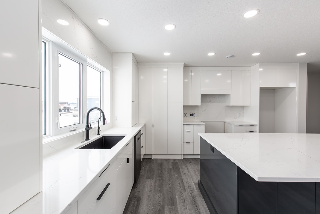 Modern kitchen with sleek white cabinetry, dark floor, and marble countertops. Large windows and bright lights create a spacious, clean ambiance.