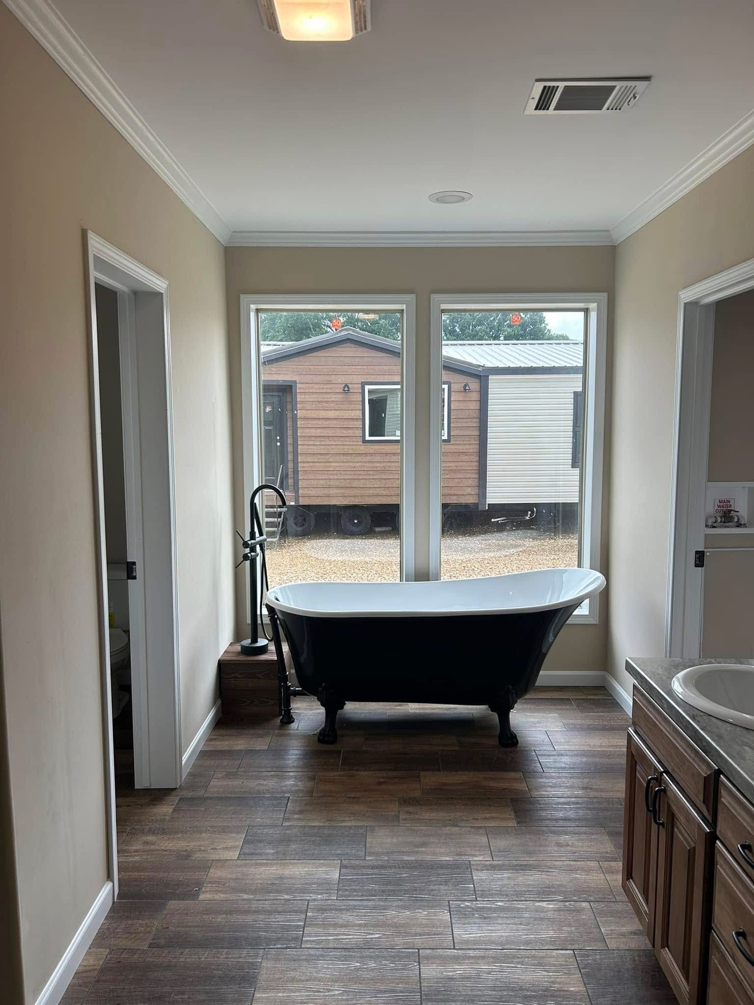 Bright bathroom with a black freestanding bathtub before large windows, revealing a view of a wooden cabin outside. Sleek, modern, serene ambiance.