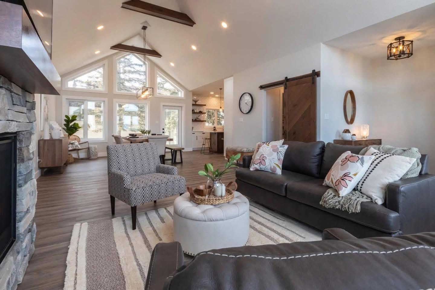 Bright, modern living room featuring vaulted ceilings, wood beams, and large windows. Cozy seating includes a dark sofa and patterned chair.