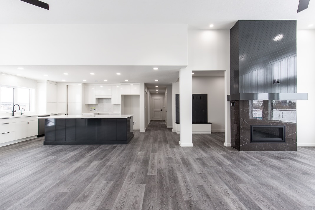 Spacious modern living area with gray wood flooring, sleek black and white kitchen, large island, marble fireplace, and ample natural light.