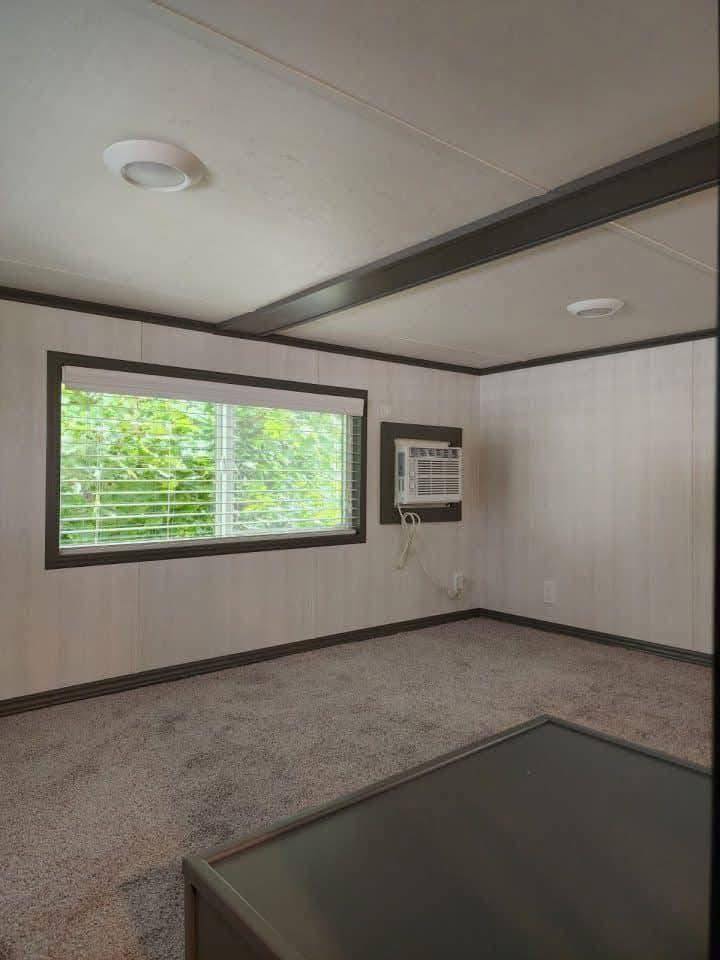 Small, empty room with gray carpet and white walls. A vintage air conditioner is installed on the wall. A large window overlooks green foliage.