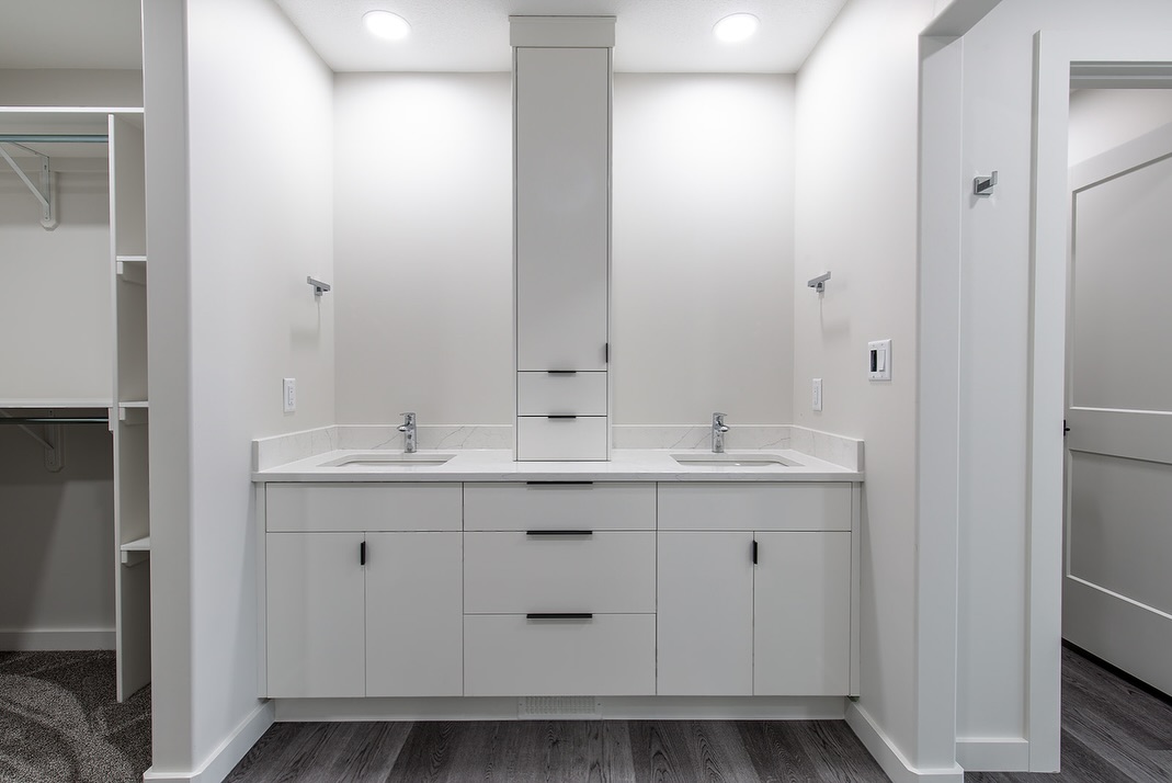 Modern bathroom with two white sinks on a sleek vanity and a central cabinet, flanked by a closet on the left. Bright lighting creates a clean, minimalist feel.