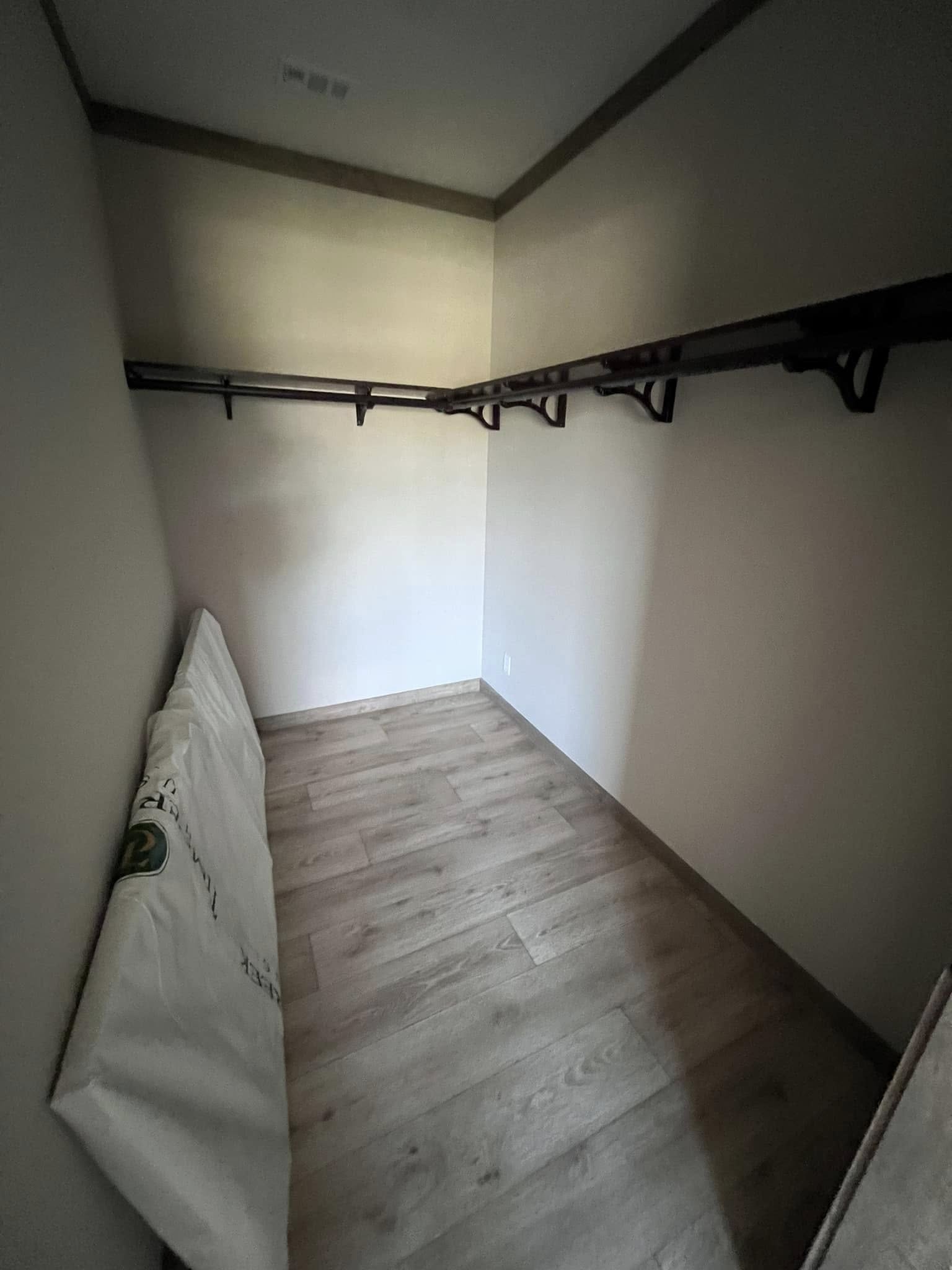 A small, dimly lit walk-in closet with wooden floors and L-shaped dark wooden shelves. A mattress is propped against the left wall. Sparse and minimalist.