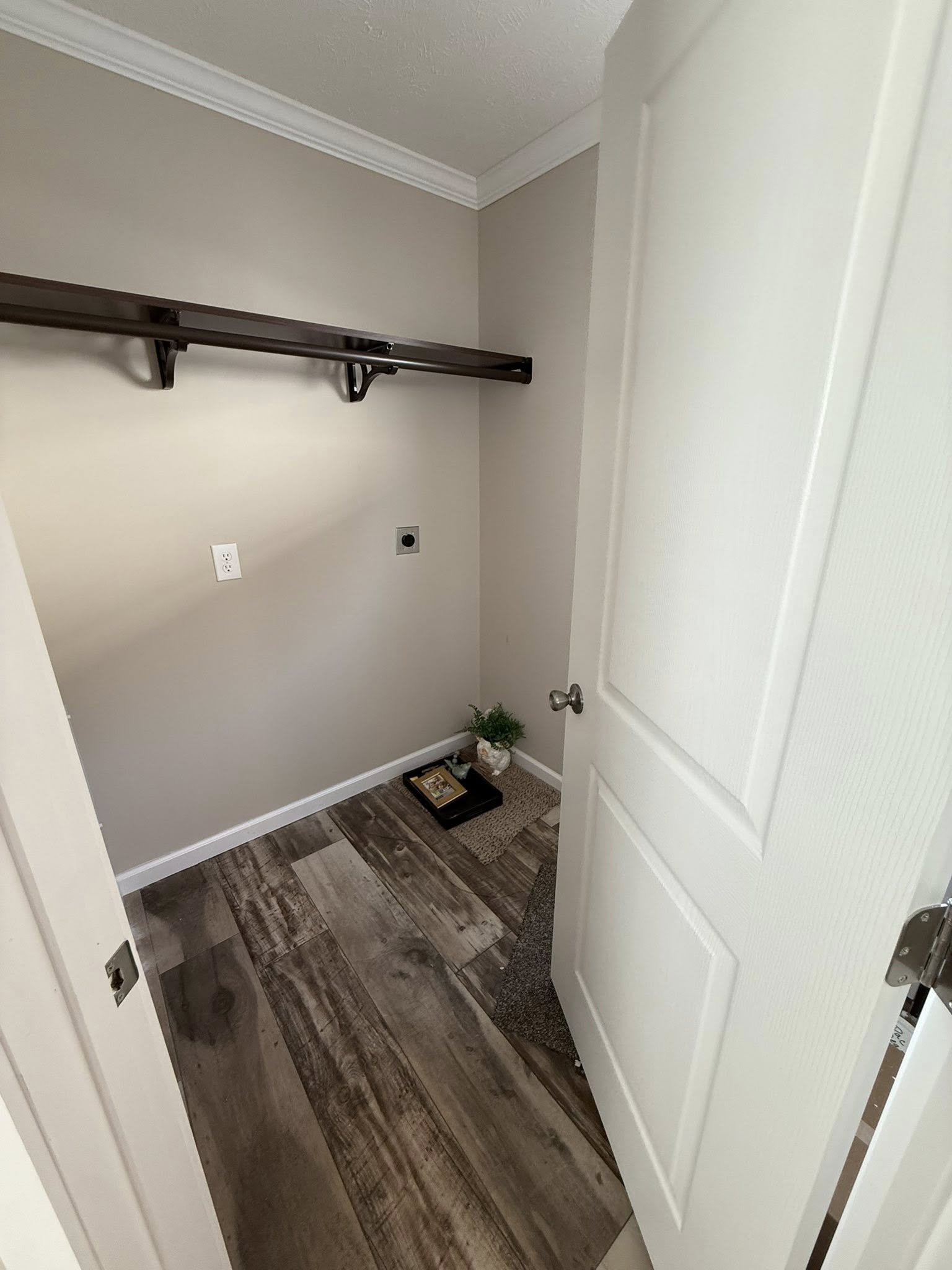 Small room with beige walls and wood flooring. It has an empty dark shelf, electrical outlet, a plant, and a small decorative item by the door. Calm and tidy atmosphere.