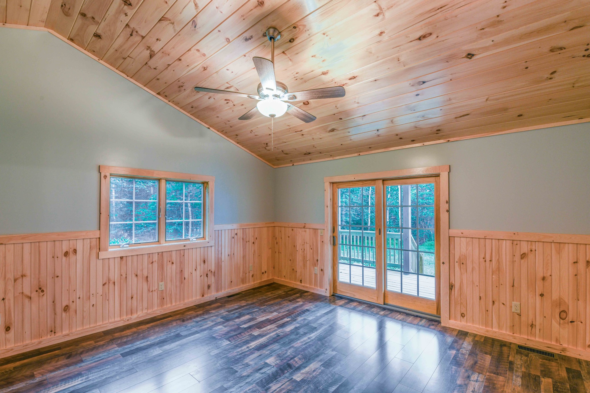 A cozy room with wooden walls and a sloped ceiling, featuring a ceiling fan. It has large windows and a door opening to a deck, with serene outdoor views.