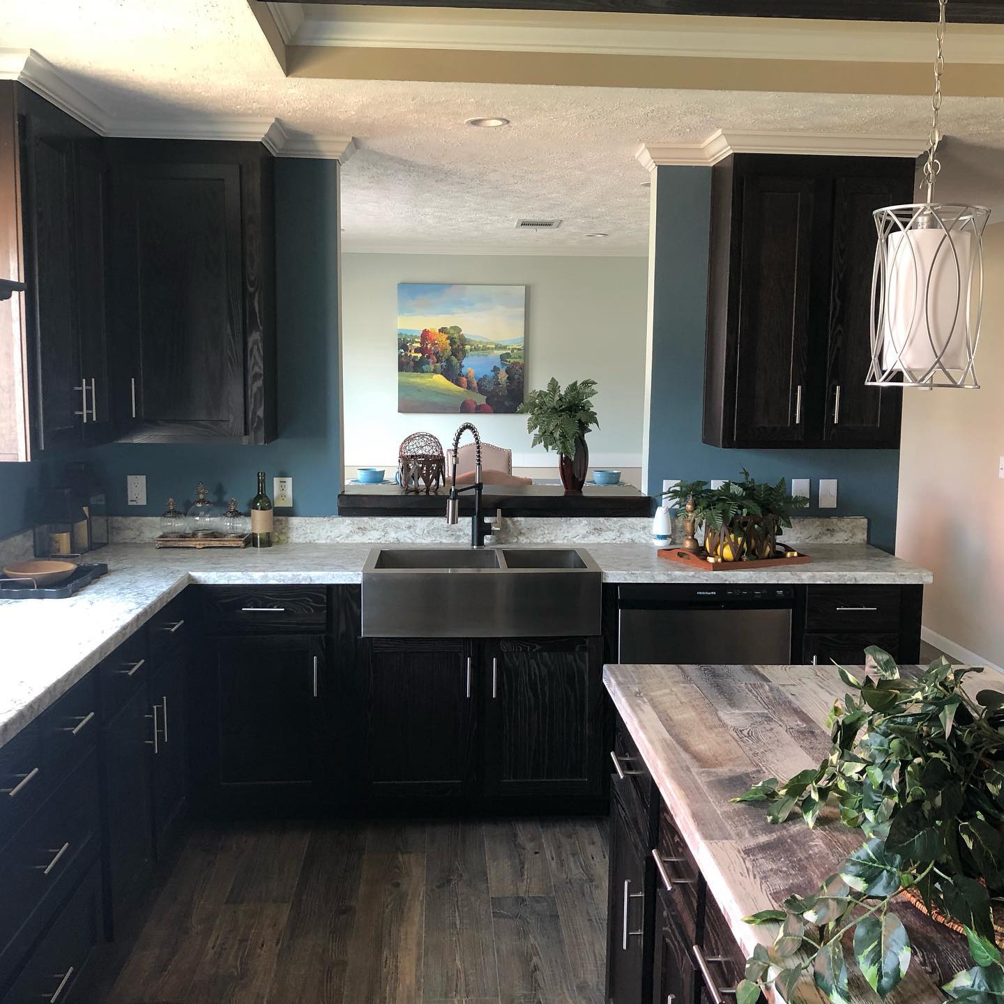 Modern kitchen with dark wood cabinets and marble countertops. A stainless steel farmhouse sink is centered below a large mirror, reflecting light. A decorative plant sits on the island, creating a cozy, inviting atmosphere.