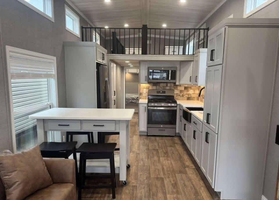 Compact modern kitchen with white cabinetry, wood flooring, and stainless steel appliances. A small island with stools and a loft area is visible. Cozy ambiance.
