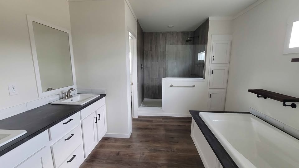 Bright, modern bathroom with white cabinets, dark countertops, and wood flooring. Features a glass-door shower and a spacious bathtub. Minimalist design.