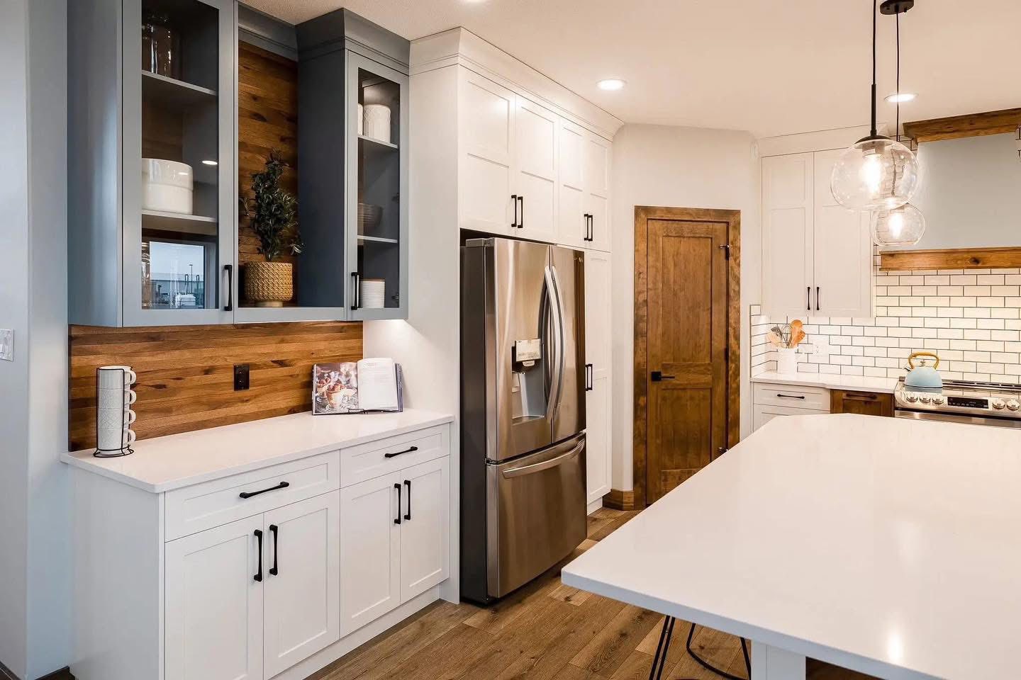 Modern kitchen with white cabinets, stainless steel fridge, and a wood-accent wall. Features a large island, pendant lights, and a warm, inviting tone.
