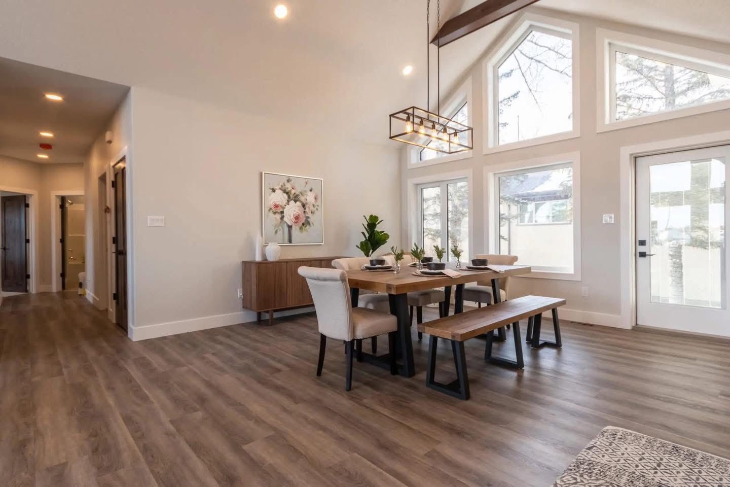 Spacious dining room with a wooden table, benches, and cushioned chairs. Large windows and modern chandelier enhance the bright, inviting atmosphere.