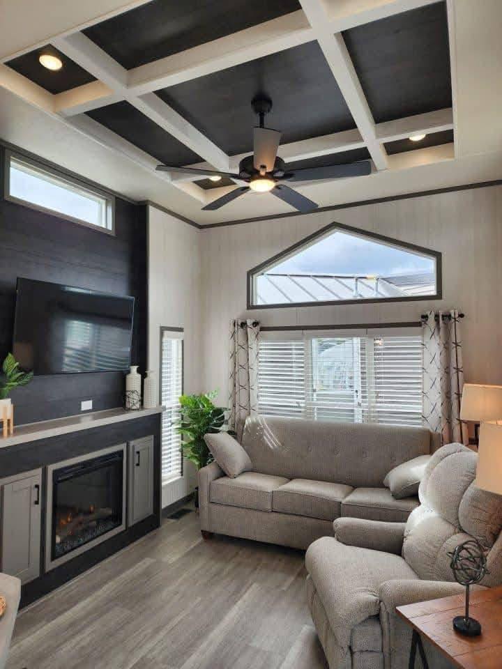 Modern living room with coffered ceiling and black fan, gray sofas, large window, and cozy fireplace. Warm tones and elegant design create a relaxing atmosphere.