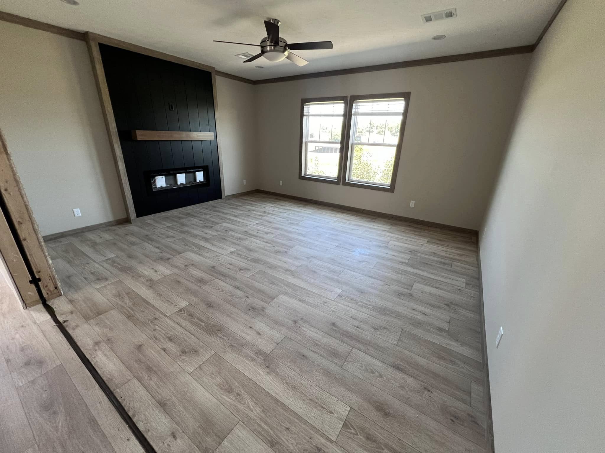 Spacious empty room with light wood flooring, beige walls, a black accent wall featuring a fireplace, ceiling fan, and two large windows with natural light.