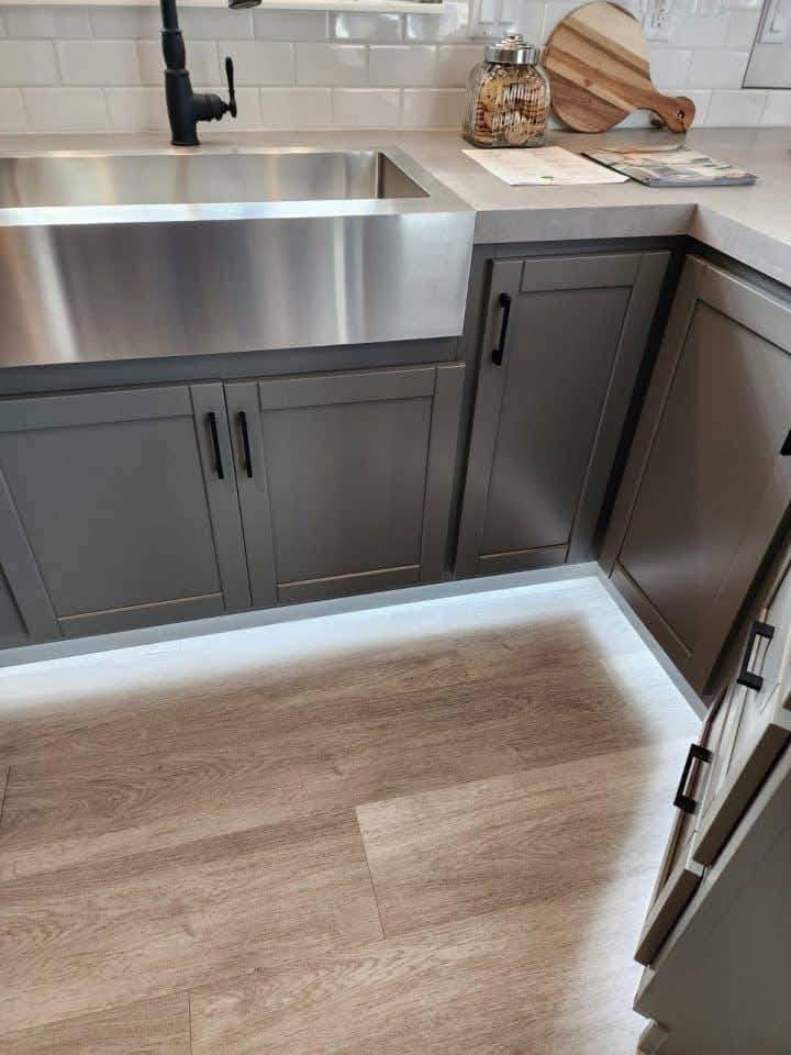Modern kitchen with a stainless steel farmhouse sink, grey cabinets, and a light wood floor. A cutting board and jar are on the counter, creating a clean and contemporary feel.