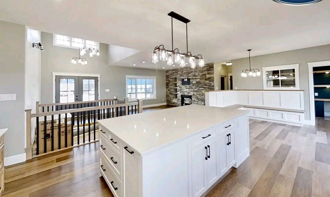 Bright open-concept kitchen and living area featuring a large white island, modern light fixtures, stone fireplace, and light wood flooring.