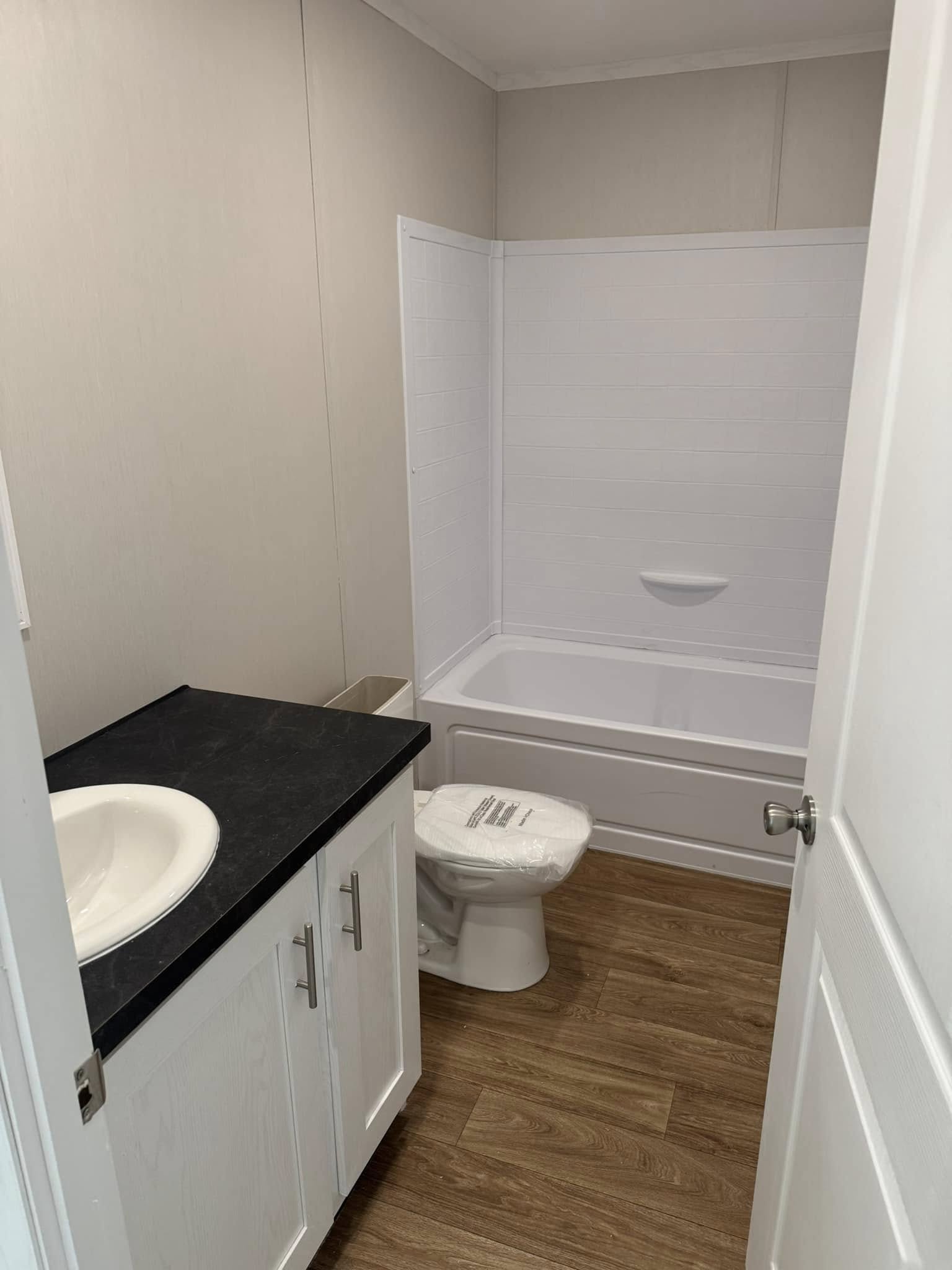 Compact bathroom with a wooden floor, white tub-shower combo, white toilet, and sink on a black countertop. Clean, simple, and functional design.
