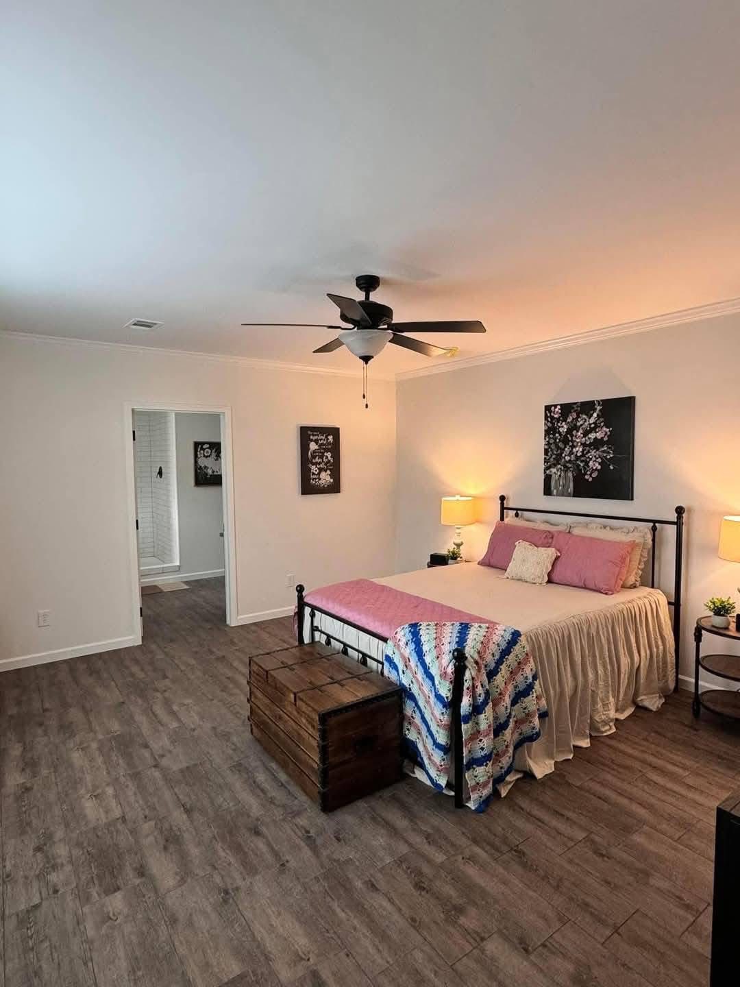 Spacious bedroom with wood flooring and a ceiling fan, featuring a bed with pink pillows, a crocheted blanket, and two side tables with lamps. Cozy, inviting atmosphere.