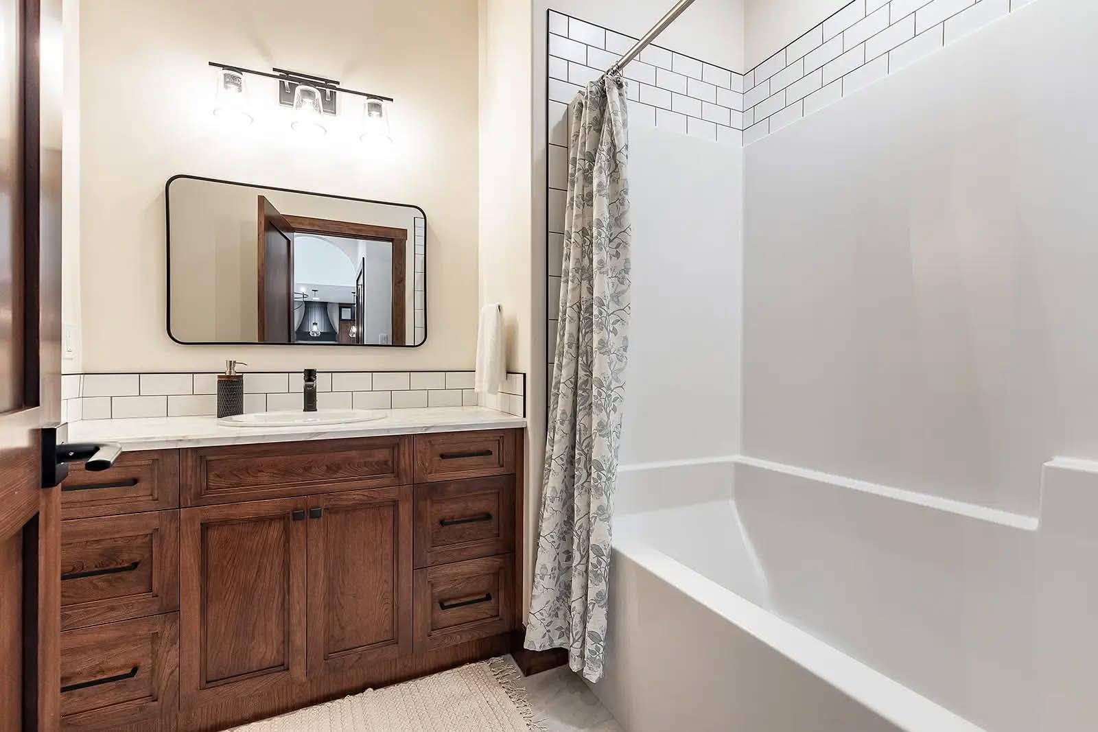 A warm, inviting bathroom featuring a wooden vanity, large mirror, and white countertop. The tub is adorned with white subway tiles and a patterned shower curtain.