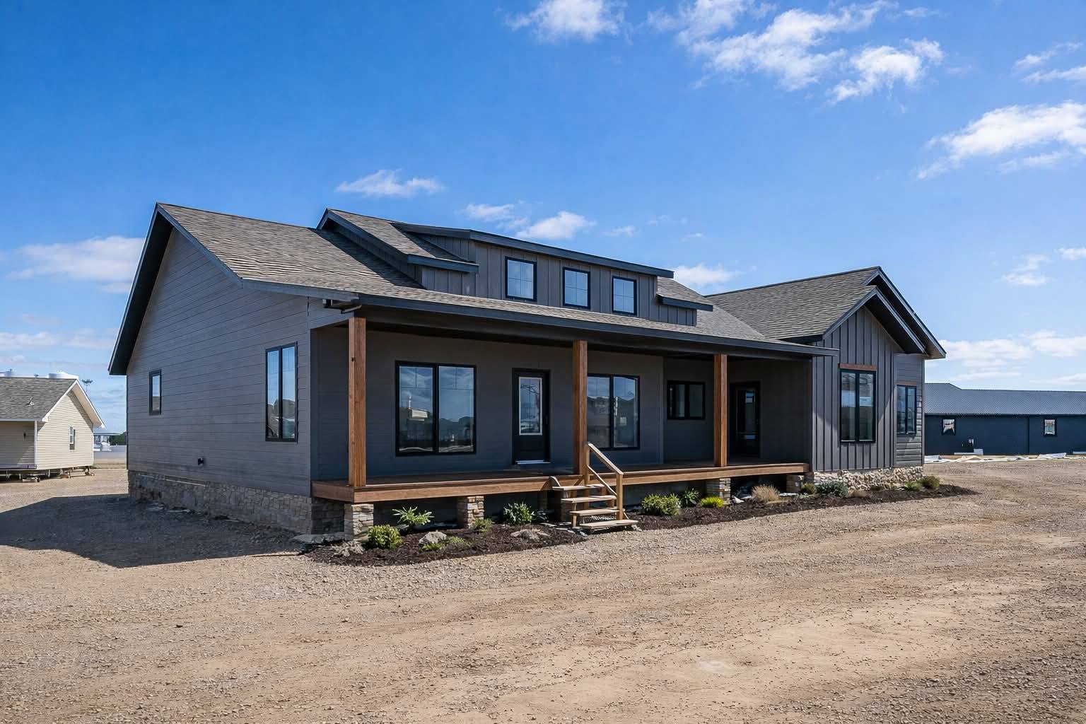 Modern single-story gray house with a large porch and wooden accents under a clear blue sky. The setting is a spacious, open area with a peaceful vibe.