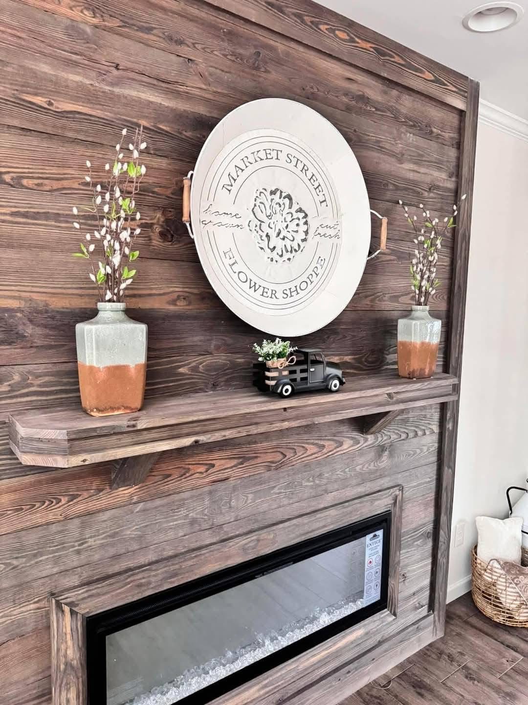 Rustic wooden wall with a circular "Market Street Flower Shoppe" sign. Below, two vases with white flowers flank a small decorative truck. Cozy and inviting.