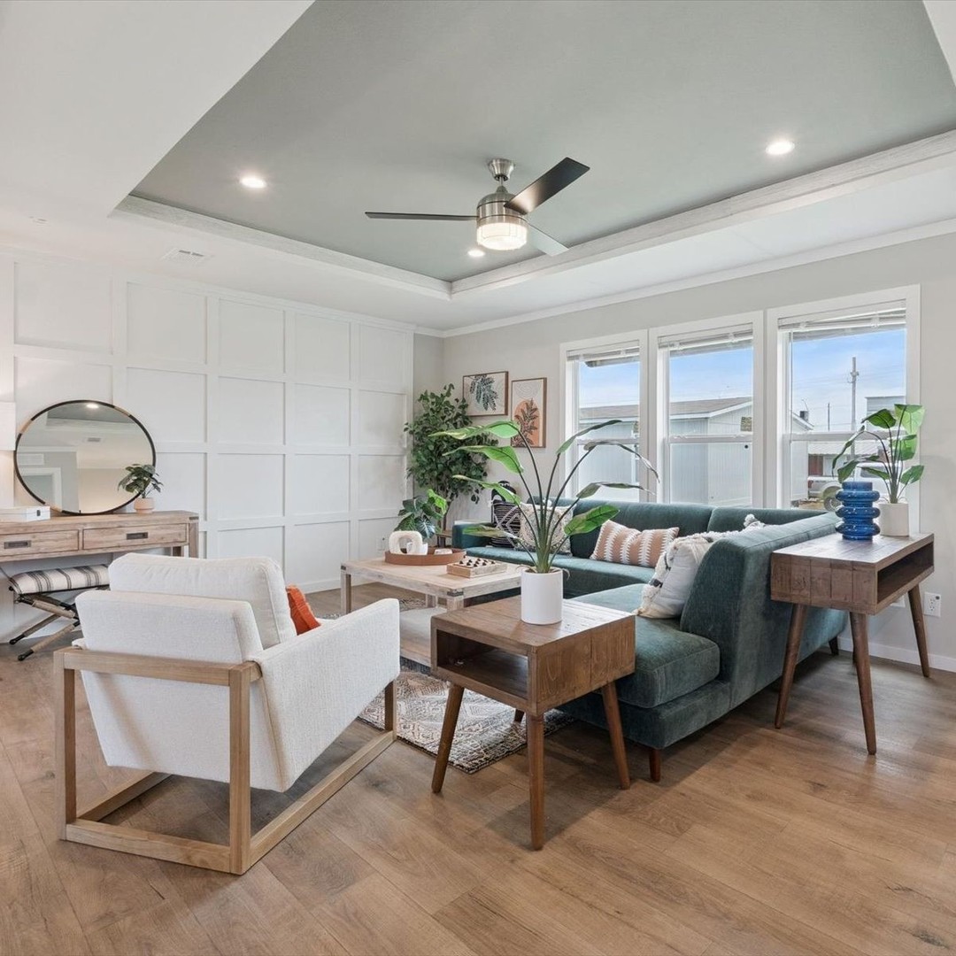 Modern living room with wood floors, teal sofa, white armchair, and assorted potted plants. A ceiling fan and large windows add light and airiness.